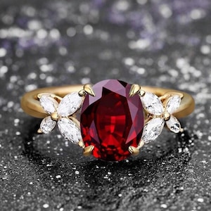 May include: A gold ring featuring a large, oval-cut, deep red gemstone as the centerpiece. Flanking the central stone are two floral clusters of white marquise-cut stones. The ring is set against a dark, textured background.