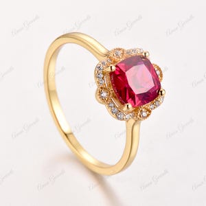 May include: A gold ring featuring a large, square-shaped, red gemstone surrounded by smaller, clear gemstones. The ring has a delicate, ornate design.
