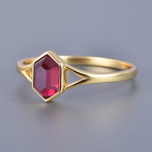 May include: A gold ring featuring a red gemstone set in a hexagonal bezel. The ring has a simple, elegant design.