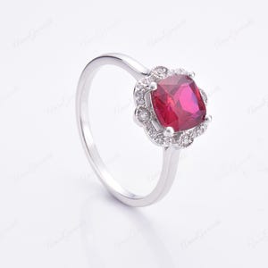 May include: A silver ring featuring a large, square-shaped red gemstone encircled by a halo of smaller white gemstones.