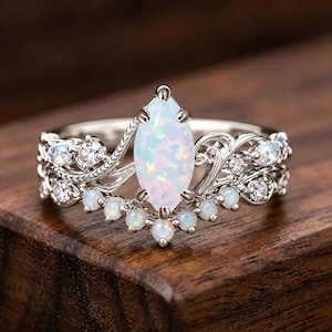 May include: A silver-toned ring featuring a central marquise-cut opal stone, surrounded by smaller round opals and clear accent stones set within a detailed metal band with swirling leaf-like patterns and prong settings throughout the design.