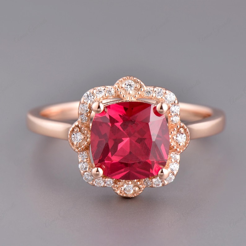 May include: A rose gold ring featuring a large, square-shaped red gemstone set within a halo of smaller, clear gemstones.