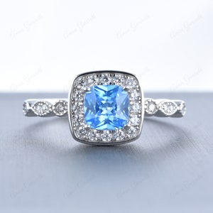 Pode incluir: Um anel de prata com uma pedra preciosa azul quadrada cercada por um halo de diamantes menores. A banda é decorada com um design delicado e intrincado.