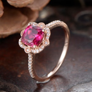 May include: A rose gold engagement ring with a cushion-cut ruby gemstone surrounded by a halo of smaller diamonds. The band is also set with diamonds.
