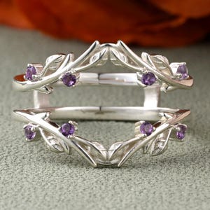 May include: A silver ring guard with a leaf design and small purple gemstones. The ring guard has a split shank design and is meant to be worn with another ring. The gemstones are evenly spaced along the leaf design.