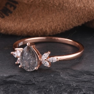 Könnte beinhalten: Verlobungsring aus Roségold mit einem birnenförmigen grauen Moissanit-Mittelstück und einem zarten floralen Design mit kleineren klaren Steinen.