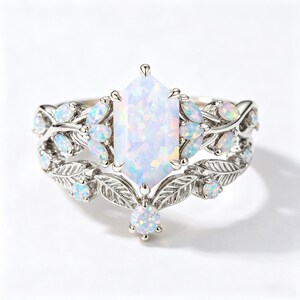 May include: A silver-toned ring featuring a central hexagonal opal stone flanked by smaller marquise and round opal accents. The band is crafted with a detailed metal leaf motif that wraps around the finger, incorporating additional small round opal stones set within the foliage design.