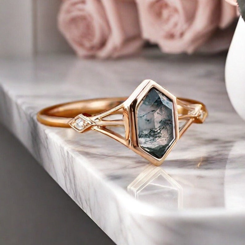 Puede incluir: Un anillo de compromiso de oro rosa con una piedra preciosa hexagonal verde y gris engastada en una banda delicada y calada.
