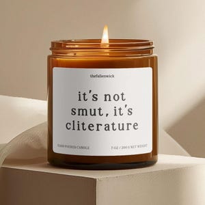 K&ouml;nnte beinhalten: Eine brennende Kerze in einem braunen Glasbeh&auml;lter mit wei&szlig;em Etikett. Auf dem Etikett steht "it's not smut, it's cliterature". Die Kerze ist handgegossen und wiegt 200 g. Der Markenname "thefallenwick" ist auf dem Etikett aufgedruckt.