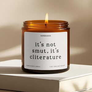 K&ouml;nnte beinhalten: Eine brennende Kerze in einem braunen Glas mit einem wei&szlig;en Etikett. Auf dem Etikett steht "it's not smut, it's cliterature". Die Kerze ist mit "Hand Poured Candle" beschriftet und wiegt 200 g netto.