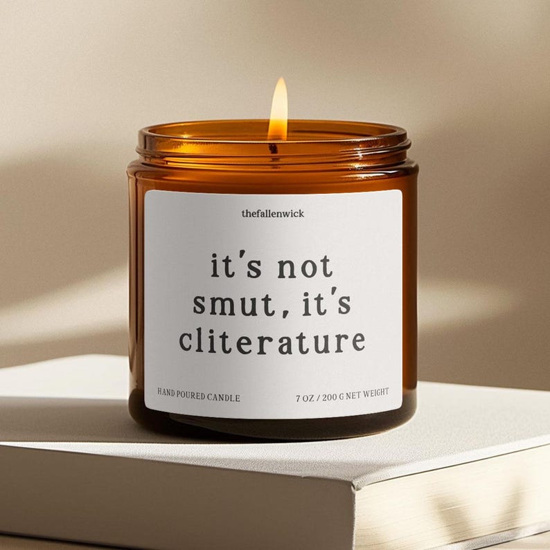 K&ouml;nnte beinhalten: Eine brennende Kerze in einem braunen Glas mit einem wei&szlig;en Etikett. Auf dem Etikett steht "it's not smut, it's cliterature". Die Kerze ist mit "Hand Poured Candle" beschriftet und wiegt 200 g netto.