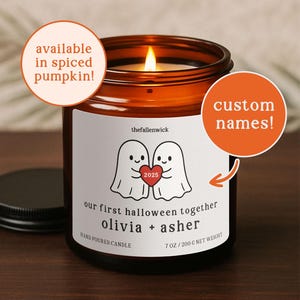 May include: A lit candle in a brown glass jar. The label shows two ghost illustrations with a heart, and the year "2025". Text on the label reads "our first halloween together olivia + asher" and "HAND POURED CANDLE". The candle weighs 200 g.