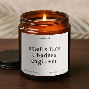 May include: A lit amber glass candle jar with a black lid. The white label reads "smells like a badass engineer." The candle is hand-poured and weighs 7 oz (200 g).