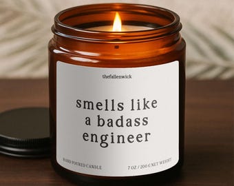 May include: A lit amber glass candle jar with a black lid. The white label reads "smells like a badass engineer." The candle is hand-poured and weighs 7 oz (200 g).