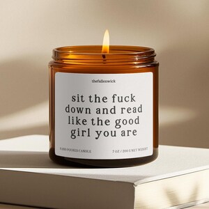 May include: A lit, amber-colored glass jar candle with a white label. The label reads "sit the fuck down and read like the good girl you are." The candle is hand-poured and weighs 7 oz (200 g).