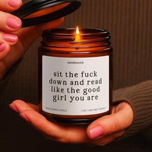 May include: A lit, hand-poured candle in a brown glass jar with a black lid. The white label reads "sit the fuck down and read like the good girl you are." The candle is 7 oz / 200 g net weight.