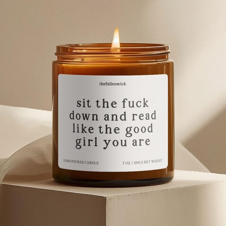 May include: A lit, amber-colored glass candle jar with a white label. The label reads "sit the fuck down and read like the good girl you are." The candle is hand-poured and weighs 7 oz (200 g).
