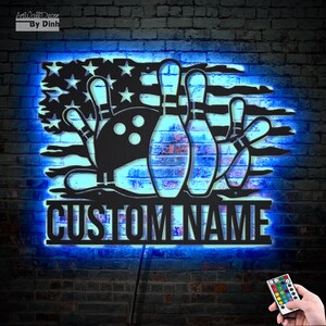 May include: Black metal wall decor featuring a bowling ball and pins with an American flag design. The decor is illuminated with blue LED lights and has a customizable nameplate that reads "CUSTOM NAME".