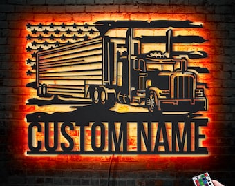 Op de afbeelding: Een zwarte metalen uitsnijding van een vrachtwagen die voor een Amerikaanse vlag rijdt. De uitsnijding is verlicht met warm oranje licht. De woorden "CUSTOM NAME" staan onder de vrachtwagen. Een afstandsbediening met meerdere gekleurde lampjes bevindt zich in de rechterbenedenhoek van de afbeelding.