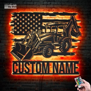 May include: Black metal silhouette of a backhoe excavator with a US flag background. The backhoe is illuminated with warm orange LED lights. The words "CUSTOM NAME" are displayed below the backhoe.