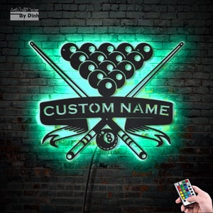 Può includere: Un decoro da parete in metallo nero con un design a stecca da biliardo e un gruppo di palle da biliardo. Il design include uno stendardo con il testo "CUSTOM NAME". Il decoro è illuminato da una luce verde brillante e viene fornito con un telecomando per cambiare il colore.