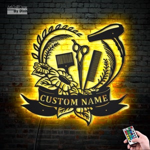 May include: Black metal wall decor with a heart-shaped design featuring a hairdryer, scissors, comb, and flowers. The text "CUSTOM NAME" is displayed on a banner below the design. The decor is illuminated with warm yellow light.