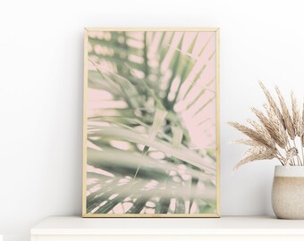 May include: A framed print featuring a soft focus image of palm fronds in shades of green and pink. The print is in a gold frame and rests on a white surface. A vase with dried grasses sits to the right.