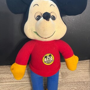 May include: A plush Mickey Mouse toy wearing a red shirt with the words "Mickey Mouse Club" on it. The toy has yellow hands and feet and blue pants.