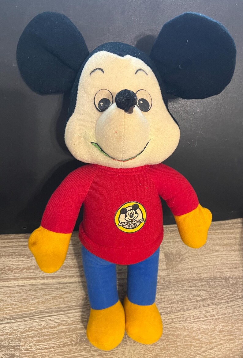 May include: A plush Mickey Mouse toy wearing a red shirt with the words "Mickey Mouse Club" on it. The toy has yellow hands and feet and blue pants.