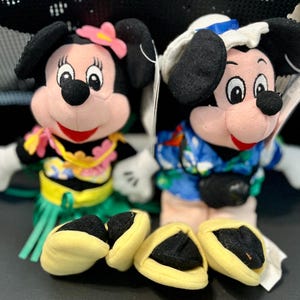 K&ouml;nnte beinhalten: Zwei Pl&uuml;sch-Mickey-Mouse-Spielzeuge, die hawaiianische Outfits tragen. Die Mickey Mouse links tr&auml;gt einen gr&uuml;nen Grasrock, eine gelbe Blumenkette und eine rosa Blume im Haar. Die Mickey Mouse rechts tr&auml;gt ein blaues und wei&szlig;es hawaiianisches Hemd und eine blaue und wei&szlig;e Matrosenm&uuml;tze.