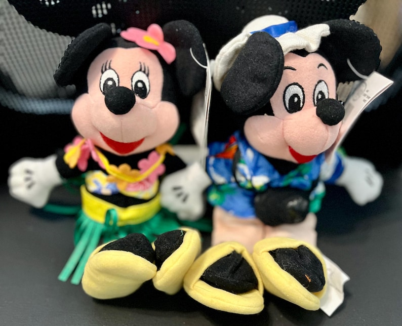 K&ouml;nnte beinhalten: Zwei Pl&uuml;sch-Mickey-Mouse-Spielzeuge, die hawaiianische Outfits tragen. Die Mickey Mouse links tr&auml;gt einen gr&uuml;nen Grasrock, eine gelbe Blumenkette und eine rosa Blume im Haar. Die Mickey Mouse rechts tr&auml;gt ein blaues und wei&szlig;es hawaiianisches Hemd und eine blaue und wei&szlig;e Matrosenm&uuml;tze.
