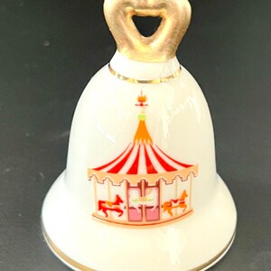 May include: A white porcelain bell featuring a colorful carousel graphic with red and white striped roof details and small horses. The bell has a gold-colored heart-shaped handle at the top and a thin gold trim around the base and the neck of the bell.