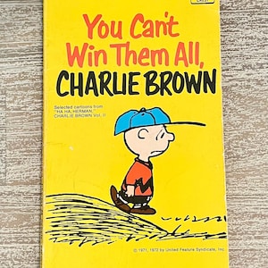 May include: A vintage paperback book titled "You Can't Win Them All, Charlie Brown." The cover is yellow with black and red text, featuring a cartoon of Charlie Brown in a blue hat and orange shirt. Published by Fawcett Crest.