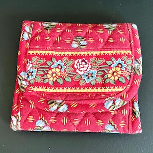 May include: A quilted fabric wallet in a vibrant red hue, featuring a repeating pattern of small blue and white flowers. A horizontal band across the center displays a detailed floral motif with red, yellow, and blue blossoms on green stems.