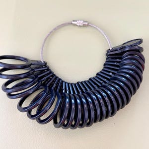 May include: A set of black plastic ring sizers arranged in a fan shape on a circular metal cable. Each individual ring sizer features a circular loop attached to a flat tab, with the entire collection held together by a silver-toned metal fastener.