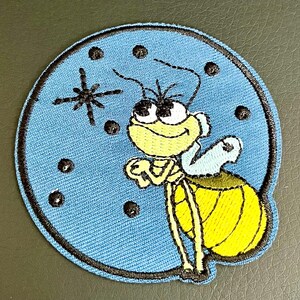 May include: A circular embroidered patch featuring a cartoon firefly with a yellow body and head, white eyes, and light blue wings. The background is light blue with black embroidered dots and a starburst design, all enclosed by a black border.