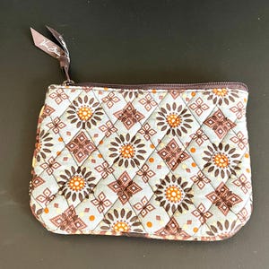 May include: A quilted fabric pouch featuring a repeating geometric pattern of brown and orange floral motifs on a light blue background. The rectangular accessory has a brown zipper closure with a fabric pull tab labeled Vera Bradley.