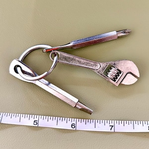 May include: A silver-toned metal keychain featuring a miniature adjustable wrench and two screwdriver bits attached to a split ring, positioned above a white measuring tape showing a scale in inches for size reference.