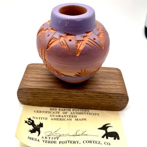 May include: A small, hand-painted pottery vase in shades of purple and orange, with carved geometric designs. The vase sits on a wooden base, accompanied by a certificate of authenticity from Red Earth Pottery, Mesa Verde Pottery, Cortez, CO.
