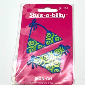 May include: A blue and green embroidered bikini iron-on patch set on a pink background. The patches feature a bikini top and bottom with a swirl design. The packaging includes the text "Style-a-bility" and "IRON-ON".