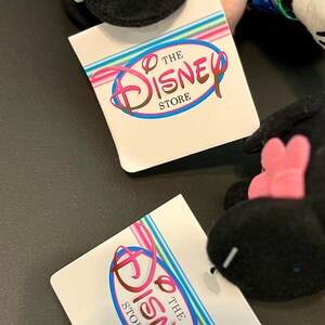 K&ouml;nnte beinhalten: Schwarz-wei&szlig;es Pl&uuml;schtier mit einem blau-wei&szlig;en Hut und einer rosa Blume. Das Spielzeug liegt auf einem wei&szlig;en Etikett mit dem Text "The Disney Store" in einem bunten Kreis.