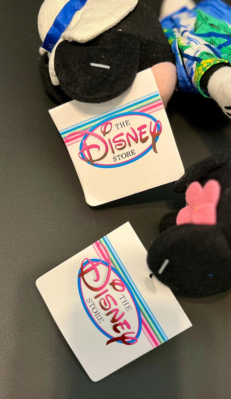 K&ouml;nnte beinhalten: Schwarz-wei&szlig;es Pl&uuml;schtier mit einem blau-wei&szlig;en Hut und einer rosa Blume. Das Spielzeug liegt auf einem wei&szlig;en Etikett mit dem Text "The Disney Store" in einem bunten Kreis.