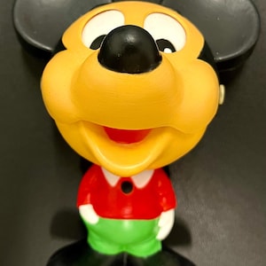 May include: A vintage Mickey Mouse figurine. The figure has a yellow face, black ears, and a black nose. It wears a red shirt, green shorts, and black shoes. The figurine is a collectible item.