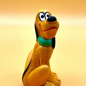 May include: A yellow Pluto figurine with black ears and a black nose. The dog is wearing a green collar and has a red mouth. The figurine is sitting down and looking up.