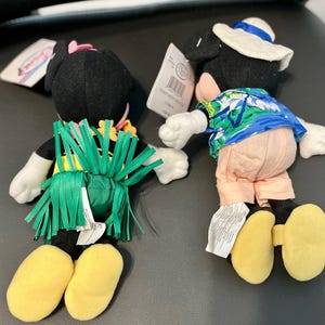 K&ouml;nnte beinhalten: Zwei Pl&uuml;sch-Mickey-Mouse-Spielzeuge, die in hawaiianischer Kleidung gekleidet sind. Ein Mickey tr&auml;gt einen gr&uuml;nen Grasrock und ein rosa Blumenband. Der andere Mickey tr&auml;gt ein blaues und gr&uuml;nes hawaiianisches Hemd und einen wei&szlig;en Hut.