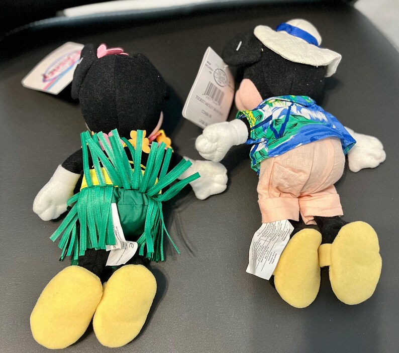 K&ouml;nnte beinhalten: Zwei Pl&uuml;sch-Mickey-Mouse-Spielzeuge, die in hawaiianischer Kleidung gekleidet sind. Ein Mickey tr&auml;gt einen gr&uuml;nen Grasrock und ein rosa Blumenband. Der andere Mickey tr&auml;gt ein blaues und gr&uuml;nes hawaiianisches Hemd und einen wei&szlig;en Hut.