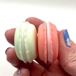 May include: Two colorful macarons, one light green and one coral pink, are held in a hand. The macarons have a white filling and a textured surface. The image is a close-up shot of the sweet treats.