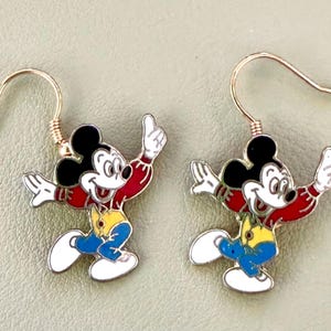 May include: A pair of dangle earrings featuring a colorful enamel charm of a cartoon mouse character. The character has black ears, a white face, a red shirt, a yellow vest, and blue pants. The charms hang from thin, curved gold-tone metal ear wires.