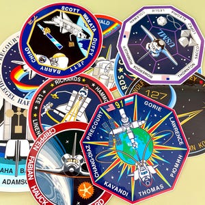 May include: A collection of circular and octagonal space mission patches featuring colorful illustrations of space shuttles, Earth, stars, and text including names like Scott, Wakata, Duffy, Jett, Barry, Chiao, Gorie, and Precourt with mission numbers.