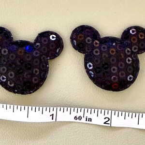 May include: Two dark purple sequined mouse-shaped patches positioned above a white measuring tape that shows the width of each patch is approximately 1.5 inches. The patches feature a circular head with two rounded ears and a dense sequin finish.